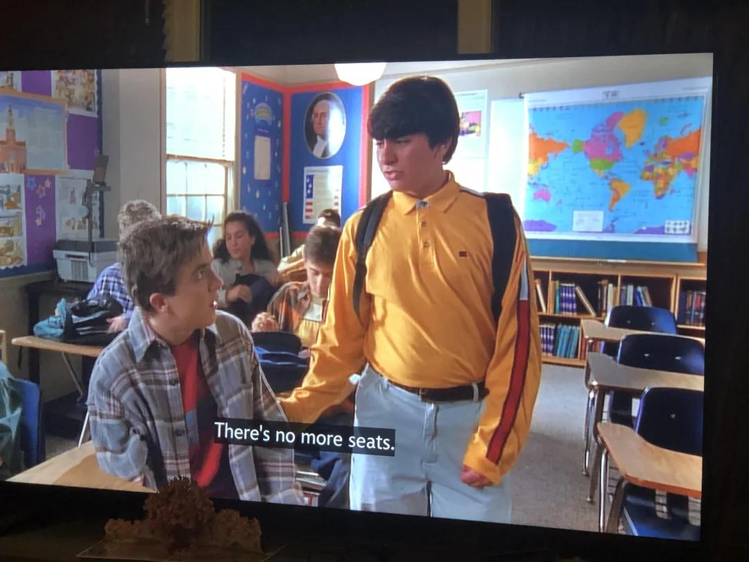 Widescreen shot of a classroom scene with one character saying they aren't any seats left while an entire empty row is visible in the shot